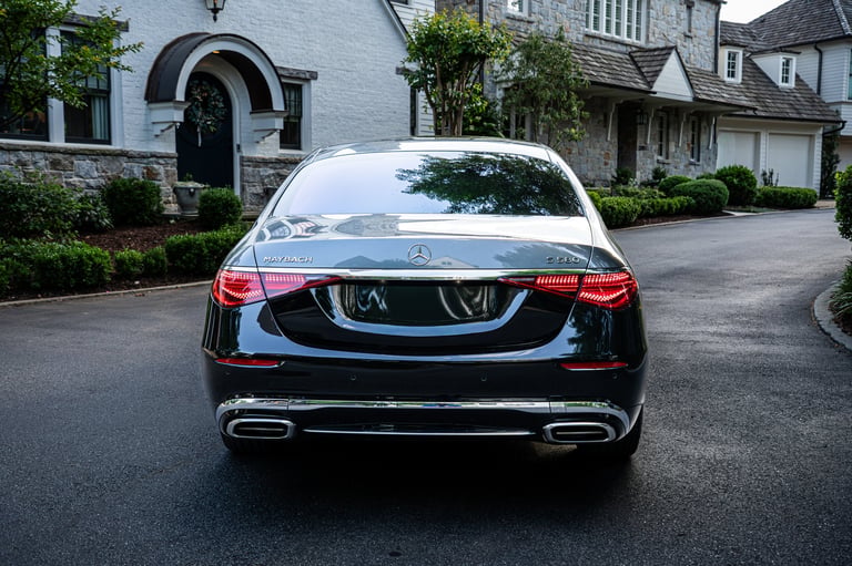 Rear view of a black Mercedes-Benz luxury sedan parked on a residential driveway with elegant white stone house in the background