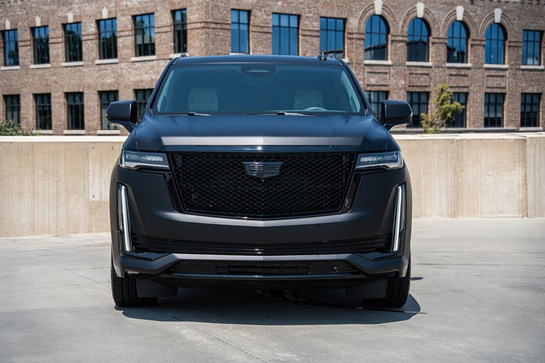 Black luxury SUV Cadillac parked on concrete lot facing camera with brick building in background