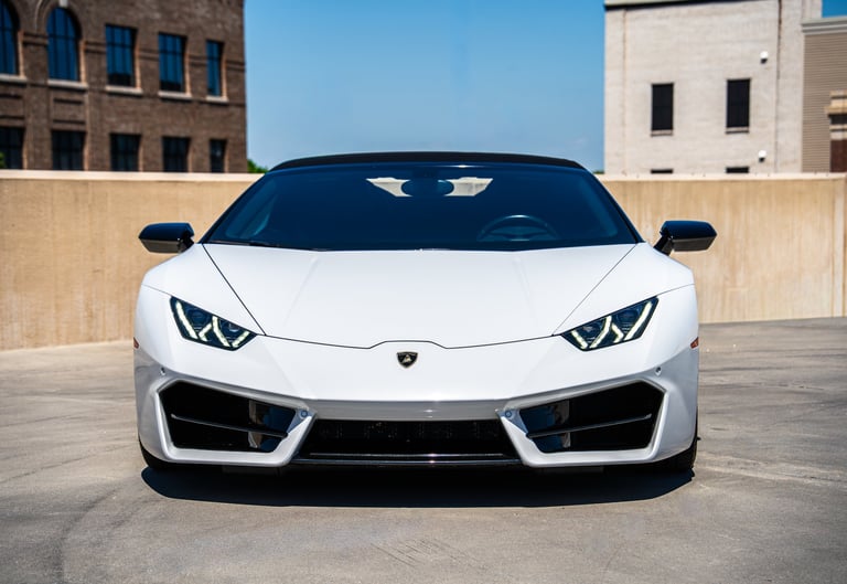 White Lamborghini Huracan sports car photographed head-on in an urban parking lot with modern buildings in the background