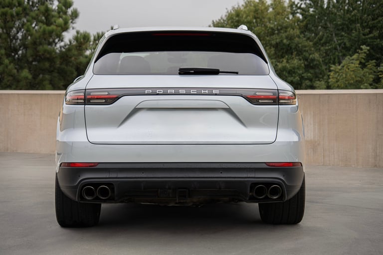 Rear view of a white Porsche Cayenne SUV with dual exhaust pipes and illuminated taillights on a concrete surface