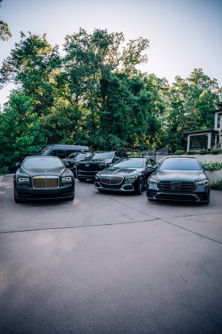 Three luxury sedans parked on a driveway in front of a home with large trees in the background