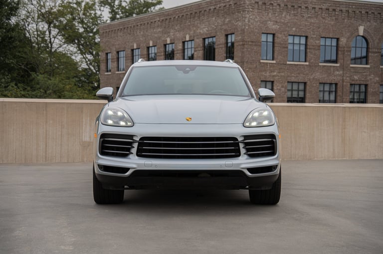White Porsche SUV viewed head-on in a parking lot with brick building and wooden fence in background