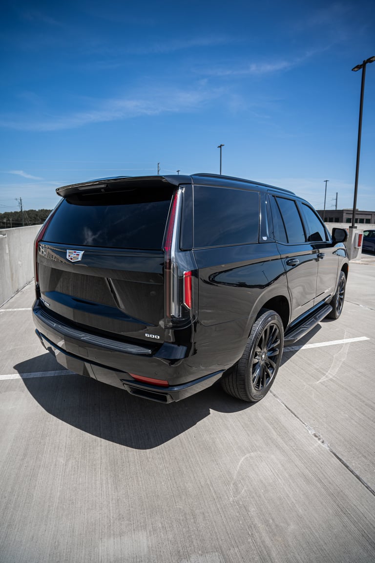 Black Cadillac Escalade SUV parked on a concrete lot under blue sky, rear three-quarter view