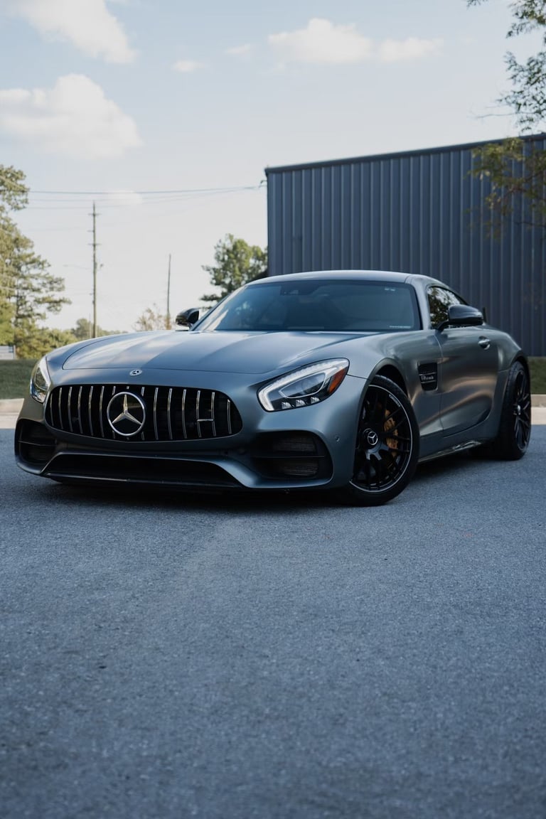 Silver Mercedes-AMG GT sports car parked on asphalt with industrial building and trees in background